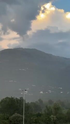 This vertical video captures a scenic view of a mountainous landscape in Dub, Montenegro. The dominant subjects are the majestic, mist-shrouded mountains in the background, dotted with small buildings, suggesting a village or scattered homes. In the foreground, a highway runs horizontally across the frame, with several cars driving on it. Green trees and vegetation line the highway and extend up the lower slopes of the mountains, creating a layered natural scene. A tall street lamp stands prominently on the left side of the highway.

The weather appears to be overcast with dramatic clouds, some of which are illuminated by the sun breaking through, suggesting either sunrise or sunset. There are subtle hints of rain or mist in the air, contributing to the hazy atmosphere. The overall mood is serene and somewhat moody, with the natural beauty of the mountains contrasting with the man-made highway and vehicles.

The time of day is likely early morning or late evening due to the lighting conditions and the dramatic cloud formations. No text is visible in the video. The activity is simply the movement of vehicles on the highway, set against the backdrop of a picturesque natural environment.
