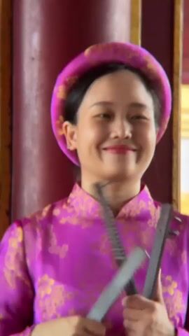 A woman in a vibrant pink áo dài with gold floral embroidery smiles as she holds two black, serrated objects. She wears a matching pink headpiece. The background shows a blurred image of a red pillar and a golden architectural element. The woman moves the objects rhythmically, creating a percussive sound as they strike each other. The clip appears to be a short, close-up shot focused on her performance.