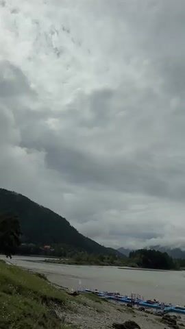 The video shows a wide, calm river flowing through a valley. The river is a muddy brown color and has several blue and pink floating structures docked along the shore. The riverbanks are covered with grey gravel on the left and green grass and trees on the right. In the background, there are lush green mountains covered in dense forests. The sky is overcast with dramatic grey clouds, giving the scene a moody and serene atmosphere. The camera pans slowly to the right, revealing a wooden footbridge extending across a small stream near the riverbank, with two people visible on it, one wearing a yellow vest.