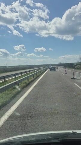 The video shows a highway with traffic moving at a moderate speed on a sunny day. The road is divided by a guardrail, and there are traffic cones in the right lane indicating a work zone. The sky is blue with puffy white clouds. There is flat, green land on either side of the highway.