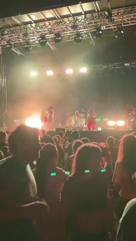 A band performs on a stage bathed in bright lights and smoke. The audience, a crowd of people, is visible in the foreground, facing the stage. Some members of the audience are holding up their phones, presumably to record the performance. The stage has a drum set and multiple musicians playing instruments. The lighting is dramatic, with spotlights and colored lights illuminating the performers. The overall atmosphere is that of a live music concert.