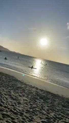 A video shows a beach scene at sunset with people on paddleboards and in a kayak. The sun is visible in the sky, reflecting brightly on the water. The beach sand is dark and textured. In the water, several people are silhouetted against the sunlight. One person is in a kayak, and others are standing on paddleboards. The video appears to have been shot from the beach, looking out towards the water.