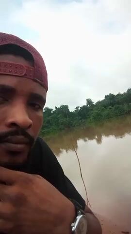 A close-up selfie video begins showing a man wearing a red baseball cap and black t-shirt. He is positioned on the left side of the frame, his face turned towards the camera. Behind him, the video pans to reveal a wide body of murky brown water, likely a river or lake. The opposite bank is densely covered with lush green trees. The sky is overcast with grey clouds. Small ripples are visible on the surface of the water in the mid-ground, suggesting movement beneath the surface. The camera continues to pan across the water, showcasing the serene yet slightly ominous natural landscape. The man is no longer visible in the frame after the initial few seconds.