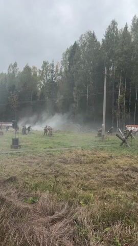 The video shows a reenactment of a battle. Several individuals dressed in military uniforms are visible in a grassy field surrounded by trees. Smoke billows from the ground in the center of the frame, indicating an explosion or a simulated one. Some individuals appear to be moving, possibly as part of the reenactment. In the background, there is a sign with red lettering on a white banner and a large red star. There are also power lines and poles in the scene. The ground is covered in dry grass. The sky is overcast. One person in the foreground is holding up a mobile phone, presumably to record the event.
