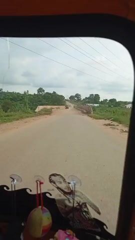 The video appears to be shot from inside a vehicle, possibly a tuk-tuk or a similar open-air vehicle, looking forward and to the sides. The first few seconds show a dusty dirt road with sparse vegetation on either side. A small bridge or overpass is visible in the distance, with another vehicle on it. There are utility poles with wires in the background. The view then shifts to the right, showing a body of water with reeds and more vegetation. The road continues, and another car is seen driving towards the camera. The vehicle appears to be moving at a moderate pace through a rural or semi-rural area. There are decorative items, possibly stickers or figurines, on the dashboard or front of the vehicle, one of which resembles a human face. The sky is overcast with some clouds.