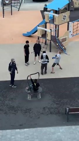 A group of children are playing on a playground. In the center of the frame, two boys are sparring with what appear to be boxing gloves. To the left of the sparring boys, two other boys stand watching. Further to the left, a boy in a black tracksuit and a grey hat stands facing the sparring boys. In the foreground, a child is sitting on a spring rider. In the background, a playground structure with slides and climbing nets is visible, and to the far left, a car is parked.