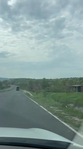 The video shows a daytime drive along a highway in Pepelishte, Bulgaria. The perspective is from inside a moving vehicle, looking forward through the windshield. The sky is overcast with scattered clouds. The highway has multiple lanes, with a guardrail separating it from the surrounding landscape. Green vegetation, including trees and grass, lines both sides of the road, and distant hills or mountains are visible on the horizon. Other vehicles, including a white van and a dark truck, can be seen ahead on the road. The weather appears bright despite the cloud cover.