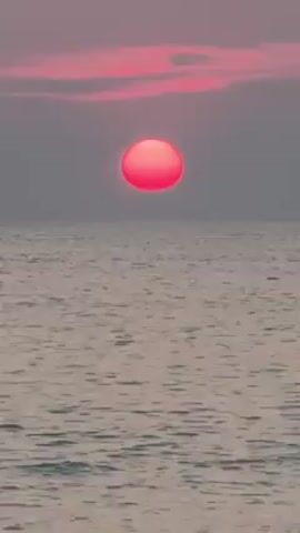 The video shows a pink sun setting over the ocean. The sky is a light gray with some pinkish clouds near the horizon. The sun is a bright pink orb, and its reflection is visible on the surface of the water. The water is calm, with small waves.