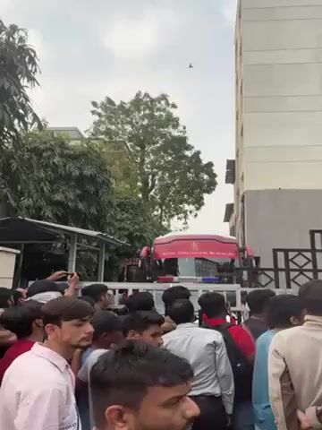 A throng of people gathers in front of the Vivanta hotel. The Royal Challengers Bangalore bus pulls up, causing excitement among the crowd, many of whom are holding up their phones to record. The building is a multi-story structure with a modern facade, and greenery, including trees and blooming bougainvillea, surrounds the area. The atmosphere appears to be charged with anticipation and excitement.