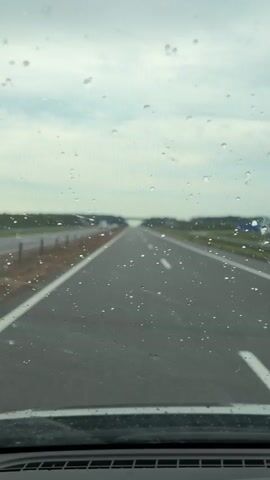 The video shows a view from inside a car, looking through the windshield, which has raindrops on it. The car is on a highway with multiple lanes. There are guardrails and vegetation along the side of the road. In the distance, a bridge is visible spanning the highway. The sky is overcast.