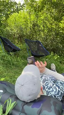 A person wearing a grey baseball cap and a t-shirt with a white and blue patterned design is lying on their back on a camouflage sleeping pad, looking at a smartphone held in their hands. They are outdoors, surrounded by lush green foliage and tall grass. In the background, there are two foldable camping chairs, one with a blue trim and the other partially visible. Two bicycles are parked nearby, equipped with saddlebags and other accessories. A small orange folding table is set up next to one of the bicycles, holding a brown paper bag, a green bottle, and other items. The overall scene depicts a moment of relaxation and preparation for outdoor activities.