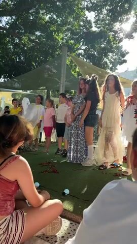 The video shows a group of children dressed in costumes standing in a line outdoors. The children appear to be participating in a performance or event. The environment is sunny with trees in the background. Some of the children are wearing animal ears or other costume accessories. There are also some adults in the background observing the children. The ground is covered with green artificial turf, and there are some decorative elements like scattered red ribbons and white balls. The camera is focused on the children, with a person in the foreground facing away from the camera. The audio includes children speaking, possibly announcing names or introducing themselves.