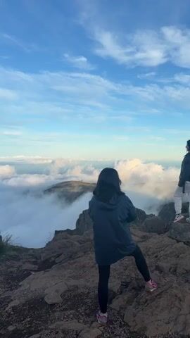 A person is standing on the edge of a rocky precipice looking out over a sea of clouds. The person is wearing a blue jacket, black leggings, and pink and grey sneakers. The sky above is blue with wispy white clouds. The ground is rocky and there is some sparse vegetation. The camera pans from the person to the clouds.