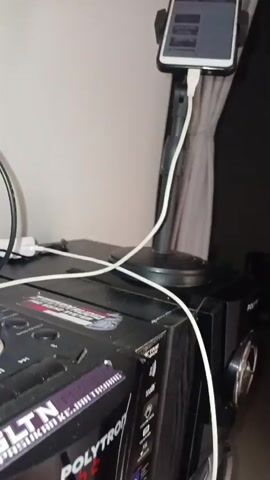 The video shows a smartphone mounted on a stand, plugged into a charger, and displayed on a table. The phone screen shows a YouTube video with a list of songs. The background appears to be a room with a wall and a curtain. The lighting is dim.