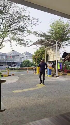 Two individuals are visible. The first, wearing a green jacket with "gojek" branding, is walking away from the camera. The second, wearing a dark blue shirt and black pants, is standing and looking towards the left. The scene appears to be an outdoor area with trees, houses in the background, and a barrier gate. The lighting suggests it is daytime.