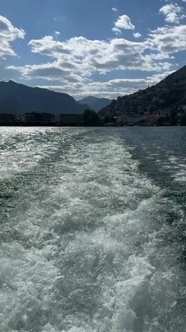 The video captures a view from a boat moving across a body of water, creating a prominent wake behind it. The water is a deep blue-green color and appears somewhat choppy. In the background, a town or city is visible built on the slopes of a large, tree-covered mountain. The architecture suggests a European setting, with buildings clustered together. The sky above is a clear blue with scattered white clouds, and the sun is shining, casting shadows. The perspective is from a low angle, looking back from the stern of the vessel. There are no people or other vessels clearly visible in the frame. The overall impression is one of scenic travel on a lake or bay on a sunny day.