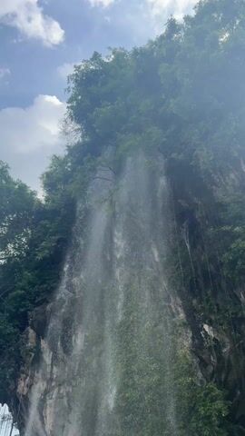 The video showcases a majestic waterfall cascading down a steep, rocky cliff face. Lush green trees and dense foliage adorn the top and sides of the cliff, with numerous vines hanging down, some almost reaching the water below. The waterfall itself is wide and powerful, creating a significant mist as it plunges into a greenish-hued body of water at its base. Several dark rocks are visible within the water near where the waterfall impacts.

The sky above is partly cloudy with patches of blue, suggesting a bright daytime setting. The camera slowly pans downwards, starting from a high perspective of the waterfall and the treeline, eventually revealing the full height of the fall and the tranquil water at its base, including the surrounding greenery. The sound of rushing water is prominent throughout the video, enhancing the immersive experience of the natural scene. The overall impression is one of serene natural beauty and the power of water.