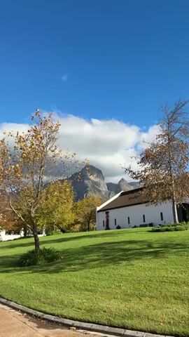 The video shows a landscape with a white building on the right, trees in the foreground, and a mountain range in the background. The sky is blue with some clouds. The grass in front of the building is green and well-maintained. Some of the trees have yellow and brown leaves. A vine with reddish leaves climbs up the side of the building. There is no visible human activity. The scene appears to be in a rural or countryside setting.