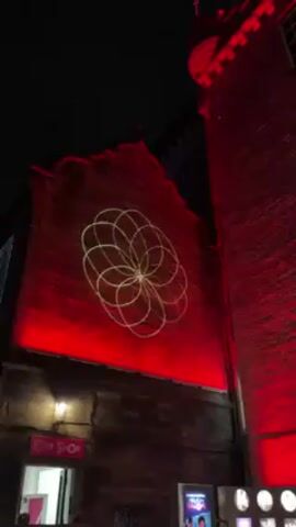 At night, two brick buildings are brightly illuminated in red. On the face of the building to the left, a white geometric design has been projected. There is a gift shop visible near the bottom of the frame. To the right, the other building is taller, with windows. Part of the Royal Mile is visible to the right of that building.