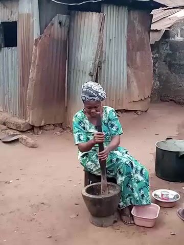 The video shows a woman sitting on a stool and using a large wooden pestle to pound something in a mortar. The woman is wearing a green and blue patterned dress and a headscarf. She looks at the camera and smiles. Behind her is a building made of corrugated metal sheets. There are a few bowls and a large pot next to her on the ground. The ground appears to be dirt.