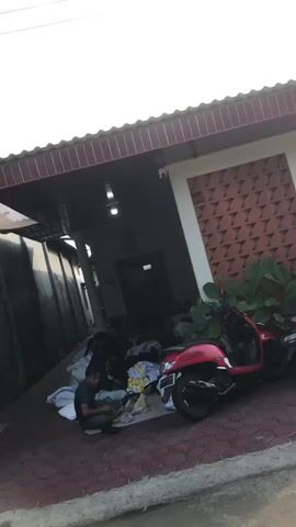 A red scooter is parked on a paved area in front of a building. Several people are gathered on the ground, some sitting and some lying down, with various items of clothing and blankets around them. The building has a covered entrance with lights and a patterned wall. The overall scene appears to be an outdoor space, possibly a driveway or a communal area.