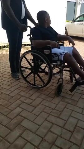 A person pushes a young person in a wheelchair on a bricked surface. The person pushing wears black slacks, a black vest, and black shoes. The person in the wheelchair wears a black shirt and white shorts. The wheelchair has black wheels and is labeled “GUARDIAN.” A white car and a white wall are visible in the background.