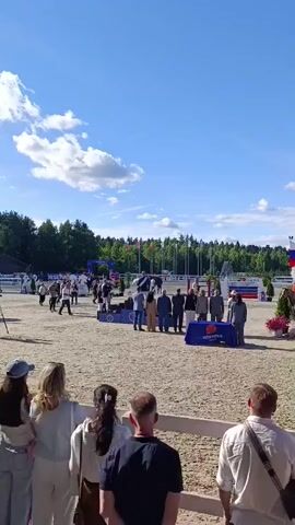 This is a vertical video capturing an awards ceremony at an equestrian event in Ivanovskoe, Russia. The scene is set outdoors on a bright, sunny day with a clear blue sky dotted with fluffy white clouds. Lush green trees form a backdrop behind a row of national flags displayed on tall poles. The ground is covered in fine, light-colored sand, typical of an arena.

In the foreground, several people are visible from behind, watching the ceremony. They are standing behind a low white fence, suggesting they are spectators. Their attire, including casual tops and pants, indicates a relaxed atmosphere.

The main focus of the video is the awards podium, where a group of individuals, likely officials and participants, are gathered. A horse and rider are on the podium, with the rider, dressed in formal equestrian attire, appearing to receive an award. Several people in suits and formal wear stand behind the podium. A blue tarp with white text, "HORSEKA," is visible on the ground in front of the podium.

The event appears to be an awards presentation following a competition. The overall mood seems celebratory and formal, with the sunshine and clear skies contributing to a pleasant outdoor atmosphere. The presence of national flags suggests an international or national-level competition. The time of day is clearly daytime, given the bright sunlight.