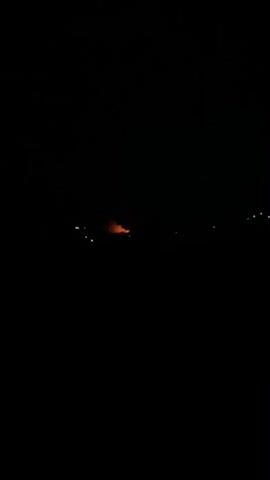 The video displays a dark night scene, with the foreground and background entirely black. In the mid-distance, a distinct orange glow, likely originating from a fire or a bright artificial light source, is visible. This central light appears to flicker or undulate slightly. Scattered across the dark expanse are several smaller, out-of-focus white lights, suggesting distant illumination from other sources or structures. No specific objects or people are clearly discernible. The scene is static, providing a view of the distant light at night in Igbara, Nigeria.