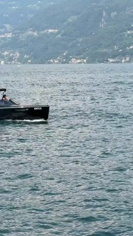 A sleek, black motorboat with a canopy glides across a calm, blue body of water. The boat is carrying a few people, one of whom is steering. In the background, a verdant, mountainous landscape rises, dotted with buildings and signs of civilization. A single sailboat can be seen in the distance. The scene appears to be set during the daytime, with clear weather, suggesting a pleasant day for boating. The number "0587CO" is visible on the side of the boat. The overall atmosphere is serene and leisurely, indicative of a vacation or recreational outing. The presence of mountains and the style of the boat hint at a European location, potentially a lake in Italy, as suggested by the provided context of Loveno, Italy.