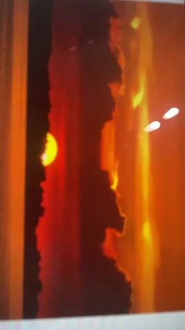 The video displays a photograph of a vibrant sunset. The sun, a bright yellow orb, is partially visible on the left side, positioned behind dark, silhouetted clouds that stretch vertically across the frame. The sky is a gradient of intense red and orange hues, suggesting the sun is low on the horizon. On the right side of the image, there are bright yellow and orange streaks, possibly reflecting light or appearing as part of the fiery sky. Two small, bright white orbs are visible in the upper right quadrant, which might be reflections or distant lights. The overall impression is one of dramatic natural beauty, with the contrast between the dark silhouettes and the brilliant colors of the sunset.