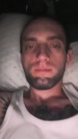 A man with a bald head, beard, and tattoos is lying in bed and flexing his bicep. He is wearing a white tank top. The video is dark and appears to be filmed at night.