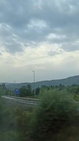 The video shows a view from a moving vehicle looking out a window. The scene is mostly out of focus due to motion blur, but it appears to be a rural or semi-rural landscape with trees, hills, and a cloudy sky. There are also some man-made structures visible, including what looks like a power line tower and a street light. The lighting suggests it might be dusk or early morning, or an overcast day.
