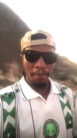 A man wearing a white polo shirt with green zig-zag patterns and the Nigeria Football Federation emblem is facing the camera. He is also wearing a beige baseball cap and black sunglasses. The background is blurred, suggesting the camera is moving or has a shallow depth of field. There appear to be natural features, possibly trees or hills, in the background. The lighting is bright, possibly due to sunlight. The man is looking directly at the camera with a neutral expression.