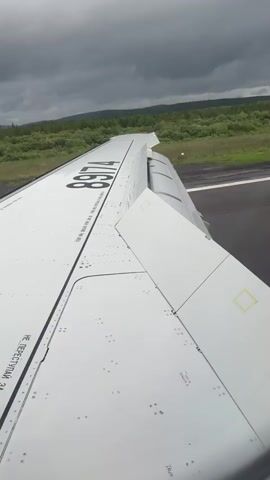 The video shows a view from an airplane window looking out at the wing and the runway below. The wing is white with black numbers and text. The runway is grey asphalt and there are green trees and hills in the background. The sky is cloudy and grey. The plane appears to be moving down the runway, possibly for takeoff or landing.