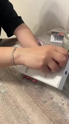The video shows a person's hands in the foreground opening a white box with an Apple logo. The box appears to be a retail package for an electronic device, likely an iPhone, given the Apple logo. The person is using their fingernails to peel off a sticker seal from the side of the box. The background shows a person wearing a black shirt and sitting at a wooden table. There is also a glass vase with a flower and some papers on the table. The lighting suggests an indoor setting.