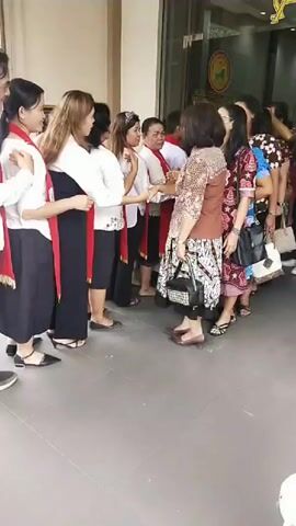 A group of people, predominantly women, are standing in a line in front of a building. Several of the women are wearing white shirts with red sashes. Some are interacting with each other, possibly in conversation or greeting. The environment appears to be an outdoor walkway or entrance to a building, with a glass door and what looks like signage visible. Other individuals are also present in the background, further down the line and inside the building. The overall impression is that of an organized gathering or event.