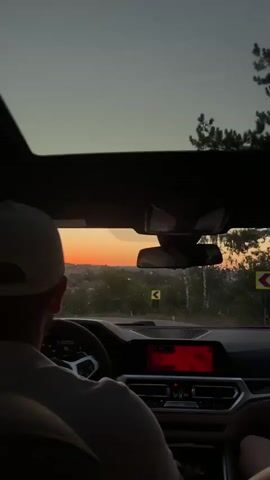 The video shows the interior of a car driving at dusk. The camera is positioned from the passenger's seat, looking forward. In the driver's seat, a person wearing a baseball cap is visible from behind, holding the steering wheel. The car has a sunroof, which is open, and a navigation screen displaying a map is visible in the center console. Outside the car, a sunset is visible over a hilly landscape with trees. The car appears to be on a paved road, and other cars can be seen in the distance. The audio features a conversation in Romanian, discussing directions and making a U-turn.