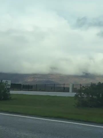 The video shows a view from a car driving along a road. On the right side of the road is a grassy area with shrubs and then a large, white building in the background. Behind the building are mountains that are partially obscured by heavy, gray clouds. The sky is overcast. The road is gray with a white line. The video appears to be taken during the day.