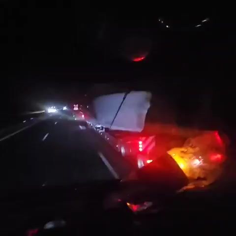 The video captures a nighttime scene from the perspective of a vehicle, likely a truck, driving on a road. Ahead, a large truck is transporting an oversized, white, tarp-covered load, which appears to be a boat or a large structural component. The rear of the transport truck is illuminated by bright red taillights and orange flashing hazard lights. Other vehicles' headlights and taillights are visible in the distance on the dark highway. Around the 0:02 mark, the camera pans to the left, briefly showing the side of the filming vehicle and the side of the oversized load. The environment remains dark throughout, indicating deep night. No people are explicitly visible, but a driver is operating the filming vehicle. There is no discernible text in the video.