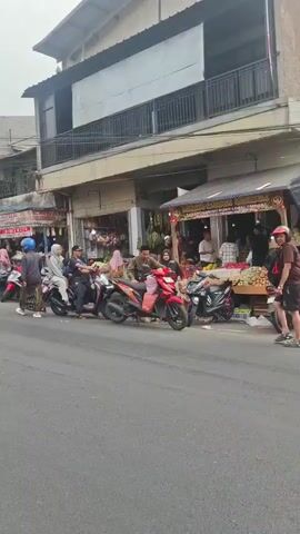 The video shows a street scene. A truck drives by. People are on motorcycles and walking. There are shops on the side of the street. The sky is overcast.