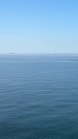 The video shows a calm blue ocean under a clear blue sky. In the distance, several ships or platforms are visible on the horizon, appearing as dark, slightly blurry shapes. The ocean surface is lightly rippled, indicating a gentle breeze. There is indistinct speech in the background.
