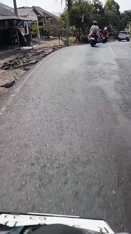 A first-person perspective video shows a person riding a motorcycle on a paved road. There are other motorcycles and a white car in front of the rider. The road is lined with trees and some buildings. There are also several flags visible on the side of the road. The rider is moving forward and the scene is slightly blurry due to the motion.