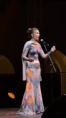 A woman is singing onstage. She is wearing a long, pale pink dress with long sleeves and gray lace detailing, and a pale gray shawl. She has light brown hair pulled back. She is standing at a microphone, gesturing with her hands as she sings. The background is dark, with a few yellow architectural elements visible, and stage lights at the bottom of the frame.