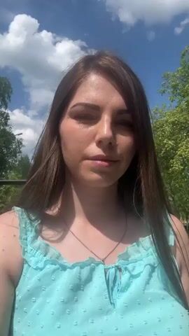 A woman stands outdoors. She is wearing a light blue sleeveless top and a silver necklace. She has long brown hair, a nose ring, and appears to be looking around. In the background there is a blue sky with some clouds, a green tree, and a metal fence-like structure.