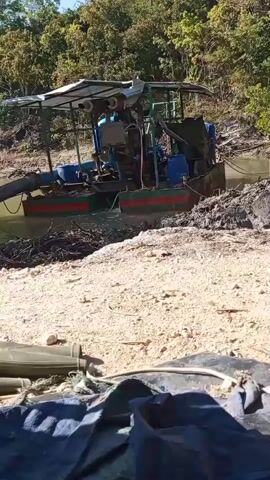 A pontoon boat equipped with a dredge pump is positioned in a body of water. A large pipe extends from the boat, submerged in the water, and then exits the water and lies on the bank. The boat has a covered canopy, a motor, and appears to be operating. The surrounding environment consists of a sandy bank with some vegetation and trees. In the foreground, a pair of green boots are laid on the ground next to a dark fabric. The water appears murky, and there is some mud or debris around the pipe where it meets the bank. The sky is clear and sunny. The video is taken from a low angle, looking up towards the boat.