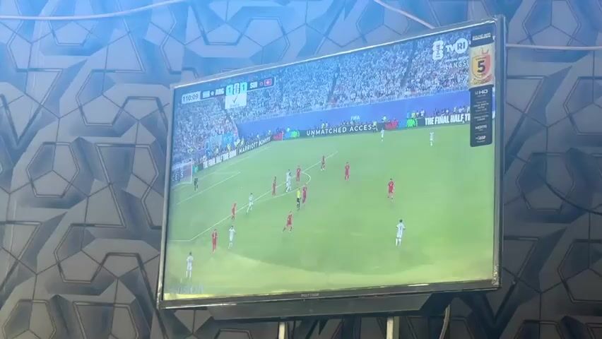 A television screen displays a soccer match in progress. The screen shows a wide shot of the field with players in red and white uniforms. The score is not clearly visible, but a timer indicates 11:03. The background of the screen shows a stadium filled with spectators. The television is mounted on a stand and is positioned in front of a wall with a geometric patterned wallpaper. The lighting in the room appears to be dim, with light shining from the top left of the frame, possibly from a projector or other light source, creating a bright, diffused illumination on the wall and partially on the television screen.