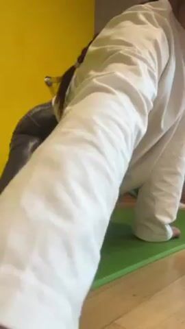 A young woman is stretching on a green yoga mat. She is wearing gray and white patterned leggings and a long-sleeved white shirt. Her hair is in a braid down her back. She starts on her hands and knees, then slowly pushes back to stretch her thighs. The background is a yellow wall.