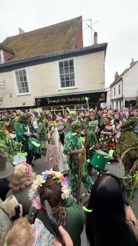 The video shows a large crowd of people gathered on a street lined with buildings. Many are dressed in green, leafy costumes, some playing drums.  A person in a light-colored dress stands out from the green costumes. Buildings on either side of the street have signs indicating businesses. Some people in the crowd hold phones up to record the parade. The weather appears overcast.