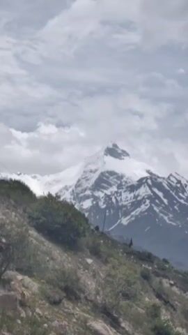 The video shows a snowy mountain peak in the distance, partially obscured by clouds. In the foreground, there is a rocky, vegetated slope. The lighting suggests it is daytime, possibly overcast. There are no people or discernible objects in the foreground besides the natural landscape. The camera appears to be static, capturing a wide shot of the mountainous terrain.