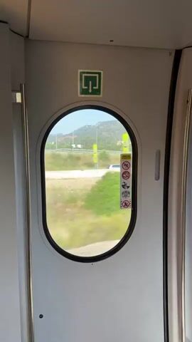 The camera is static inside a train carriage, focused on an oval window. Outside, the landscape rushes past, featuring green vegetation, roads, and occasional buildings under a blue sky. The window has a series of warning stickers attached. The train carriage's interior is white, with metal handles on the door frame. A green emergency exit sign is visible above the window.